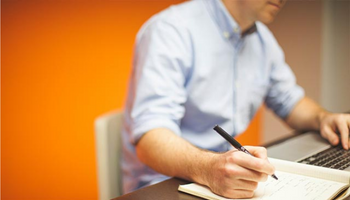 Man taking notes with pen and paper in front of a laptop