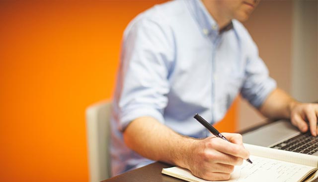 man sitting at desk with laptop writing on a piece of paper