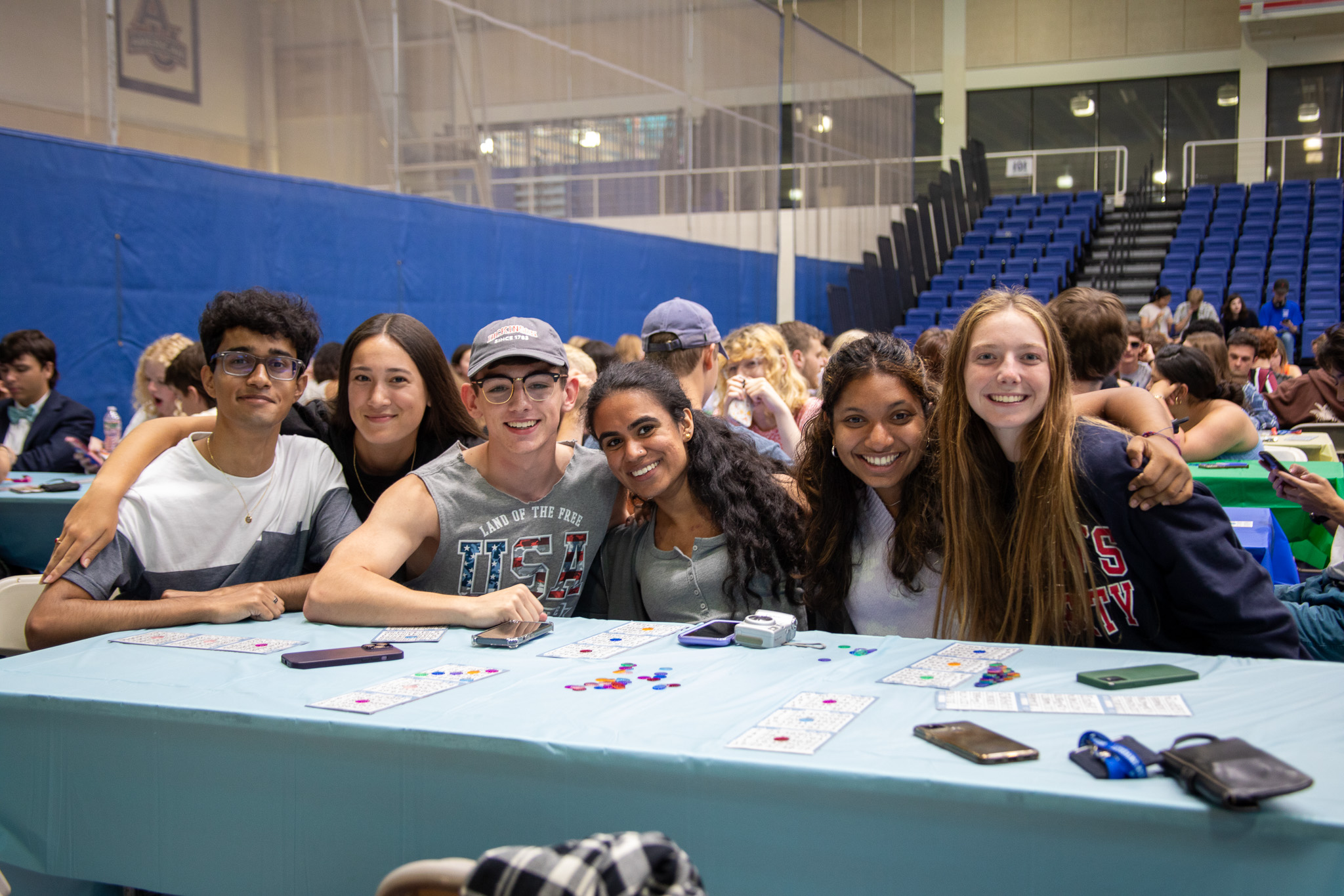 A group of students smiles at the camera, all sitting at a table in Bender Arena. They are playing bingo on the table.