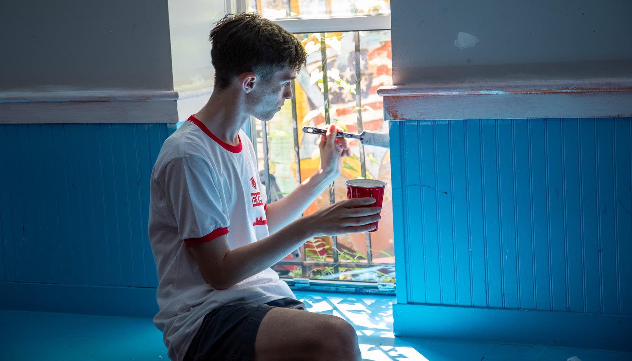 Student painting wall while seated in the window.